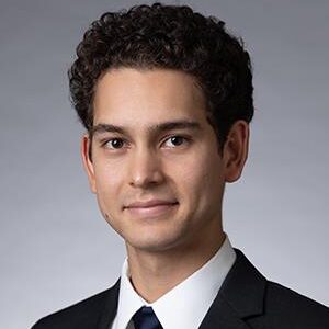 Shane, a man with black hair, wears a white shirt, black tie and black blazer.