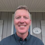 Don O'Toole is a white man with light brown hair. He is smiling, wearing a dark grey button-down shirt, and standing outside of a grey building.