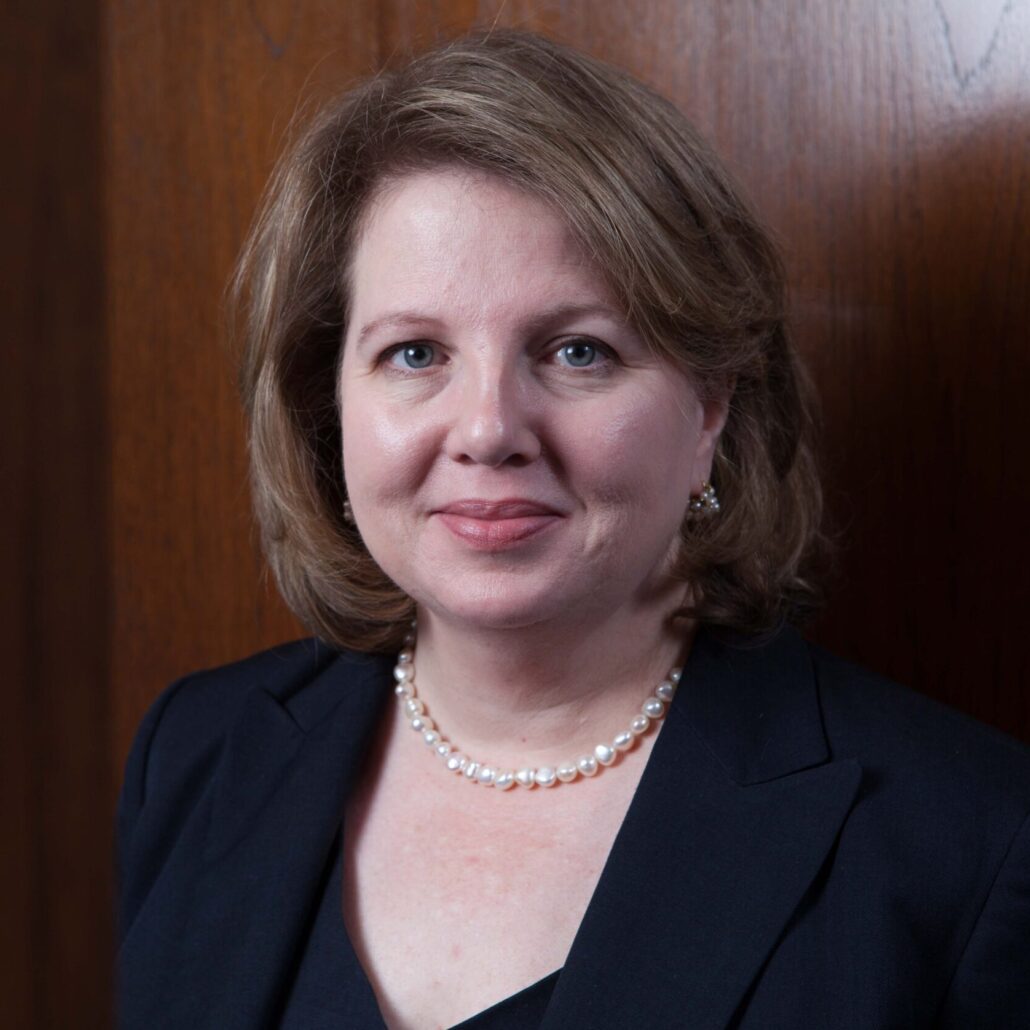 Eleanor, a white woman with brown hair, wears a black dress, black blazer, and pearl necklace.
