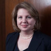 Eleanor, a white woman with brown hair, wears a black dress, black blazer, and pearl necklace.