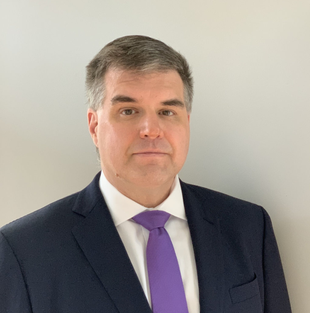 George, a white man with grey hair, wears a white shirt, purple tie and black jacket.