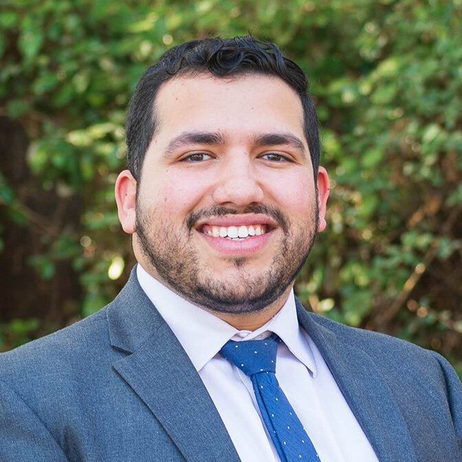 Carlo, a man with brown hair and a beard, wears a white shirt, blue tie and grey suit.
