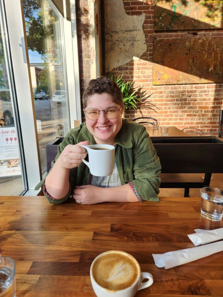 MJ, a white person with brown curly hair, wears a grey shirt and olive green button-down shirt. MJ is holding a coffee cup in a brightly lit coffee shop and sitting with a window in the background.