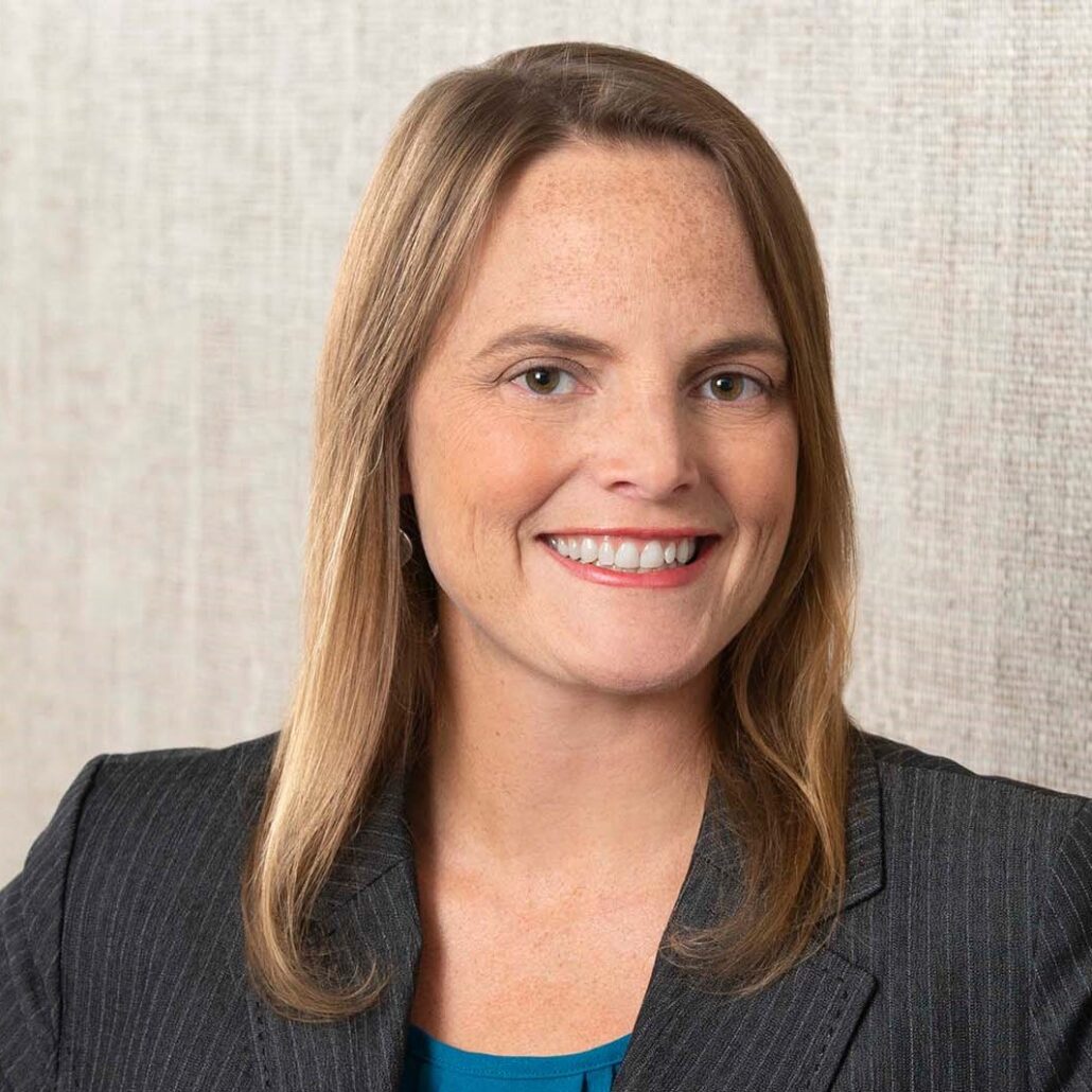 Amie, a white woman with light brown hair, wears a teal blouse and dark grey jacket.