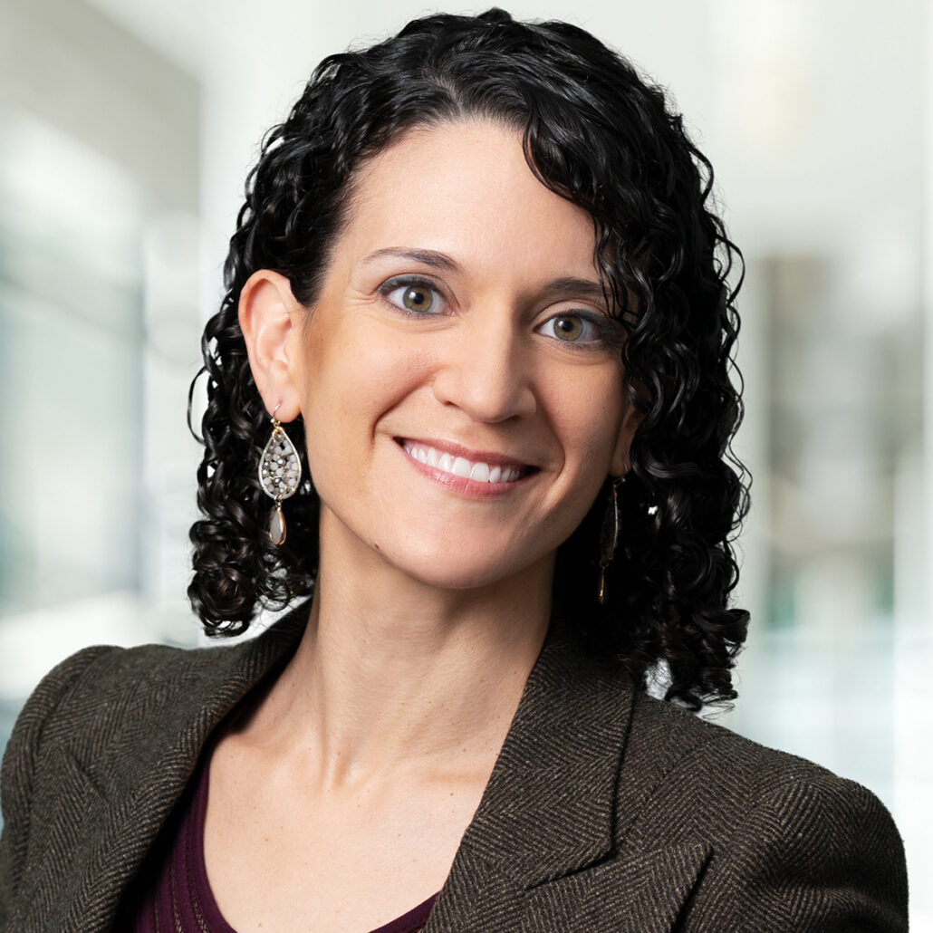 Michelle, a white woman with black curly hair, wears a dark purple blouse, dark grey jacket, and silver earrings.