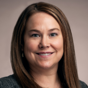 Sara, a white woman with brown hair, wears a black shirt and grey blazer.