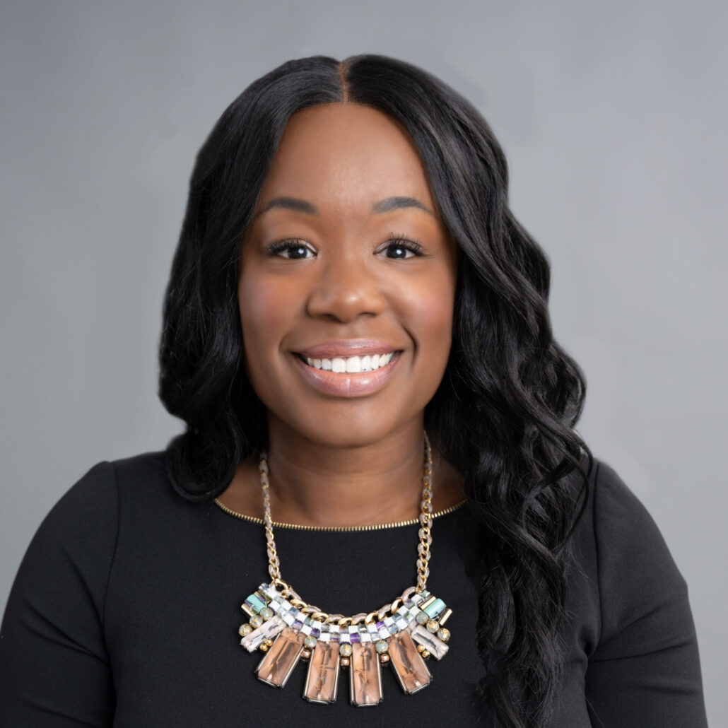 Natasha, a Black woman with black hair, wears a black blouse and gold necklace with white and peach stones.
