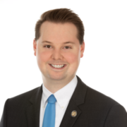 Matthew, a white man with dark brown hair, wears a white shirt, blue tie and black jacket. 