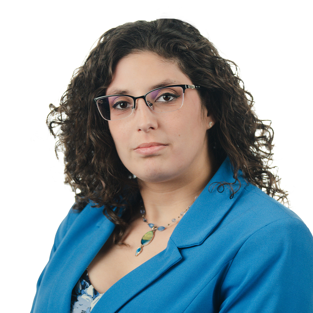 Yazmeen, a woman with curly brown hair and brown glasses, wears a blue blouse and bright blue blazer.