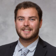 Nicholas, a white man with brown hair, wears a grey shirt, grey and white striped tie, and black blazer.