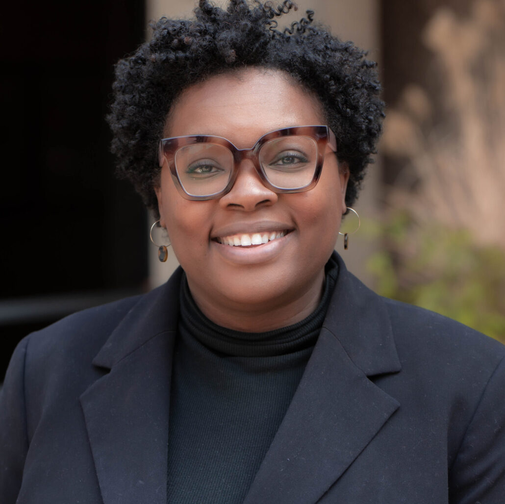 Deja, a Black woman with black hair, wears brown glasses, a black blouse and a dark grey blazer.