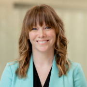 Alicia, a white woman with golden brown hair, wears a black blouse and teal blazer. 