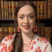 Savannah, a white woman with brown hair, wears a white sweater with coral flowers and coral earrings and a necklace.