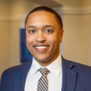 Michael, a Black man with black hair, wears a white shirt, black and off-white striped tie, and navy blazer. 