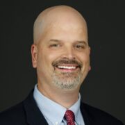 Alan, a white man with a shaved head and a brown mustache, wears a blue shirt, red tie and black jacket. 