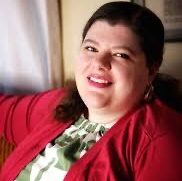 Emily, a white woman with brown hair, wears a green and white blouse and red sweater.