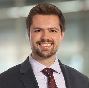 Nathan, a white man with brown hair and a beard, wears a white shirt, grey suit and burgundy tie with a white pattern.