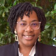 Bianca, a Black woman with black hair and clear glasses, wears a white blouse with a black print, a black blazer, and gold earrings.