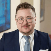 Collins, a white person with brown hair and a beard, wears a white shirt, light blue tie and navy suit.
