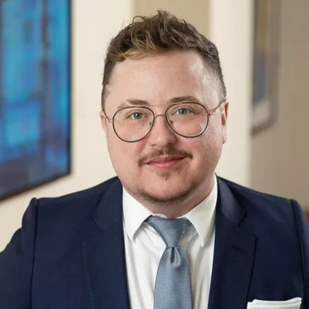 Collins, a white person with brown hair and a beard, wears a white shirt, light blue tie and navy suit.