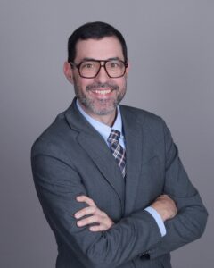 Jonathan, a white man with brown hair and brown glasses, wears a blue shirt, plaid tie and grey suit.