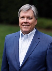 Mark, a white man with grey hair, wears a pale blue shirt and blue suit. 