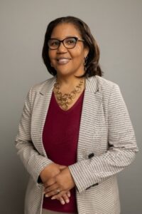 Genell, a woman with brown hair and brown glasses, wears a crimson blouse and beige jacket. 