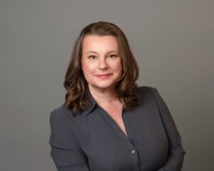 Anna, a white woman with brown hair, wears a dark grey suit. 