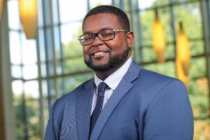 J. Hamilton is a Black man with black hair and a beard and black glasses. He wears a white shirt with a dark blue tie and a grey suit. 