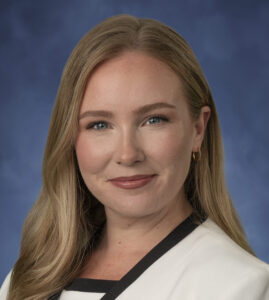 Elizabeth, a white person with blond hair, wears an off-white blouse with a black border and an off-white suit with a black border.