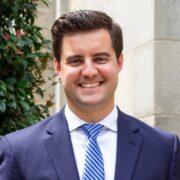 Zach, a white man with brown hair, wears a white shirt, blue tie and navy suit. 