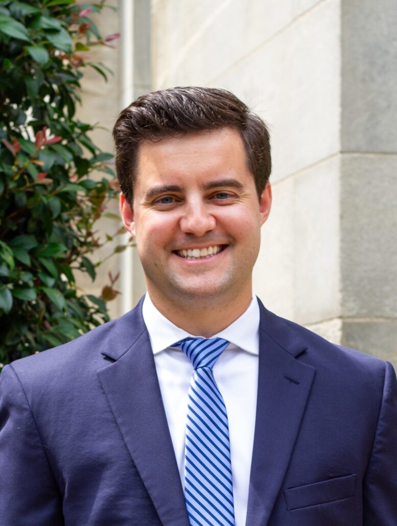 Zach, a white man with brown hair, wears a white shirt, blue tie and navy suit.