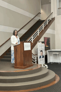 Helen is a white woman with brown hair. She wears a blue and white dress and white jacket.