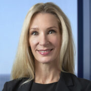 Elizabeth, a white woman with blond hair, wears a black blouse and a black jacket. 