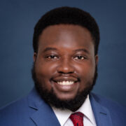 Jonathon, a Black man with black hair and a beard, wears a white shirt, red tie and blue suit.