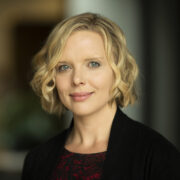 Kym, a white woman with blond hair wears a black dress and black blazer.