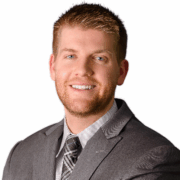 Alec, a white man with light brown hair and a beard, wears a grey shirt, grey plaid tie and dark grey suit.