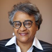 Bernice, a woman with grey hair and black glasses, wears a white shirt and black sweater.