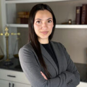 Olivia, a woman with dark brown hair, wears a black blouse and grey blazer. 