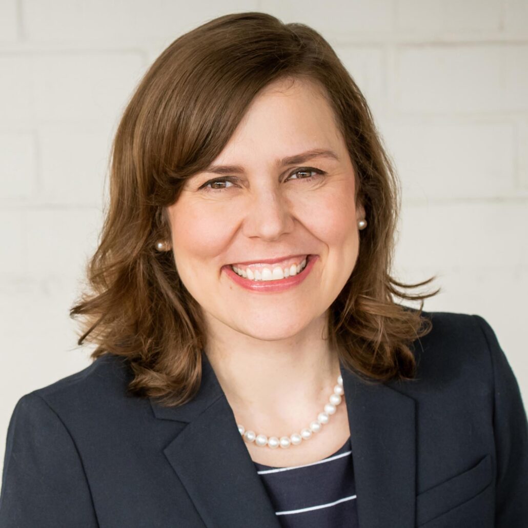 Kyla, a white woman with brown hair, wears a black blouse with white stripes and a black blazer.