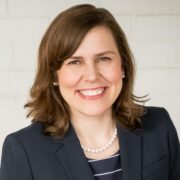 Kyla, a white woman with brown hair, wears a black blouse with white stripes and a black blazer.