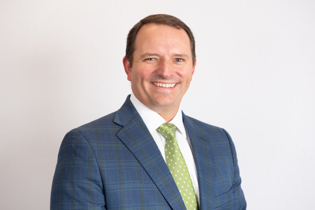 Peter, a white man with brown hair, wears a white shirt, bright green tie with white polka dots, and blue suit with light brown plaid.