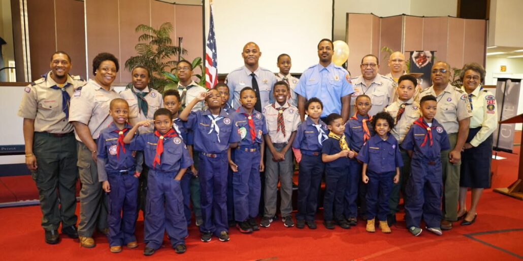 A group of volunteers and cub scouts in uniform are pictured in the lobby of a church with the American flag in the background.