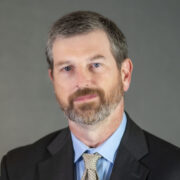 Frank, a white man with brown hair and a beard, wears a blue shirt, grey tie and black suit.