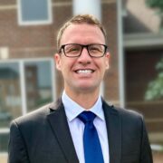 Stephen, a white man with light brown hair and black glasses, wears a pale blue shirt, blue tie, and black suit.