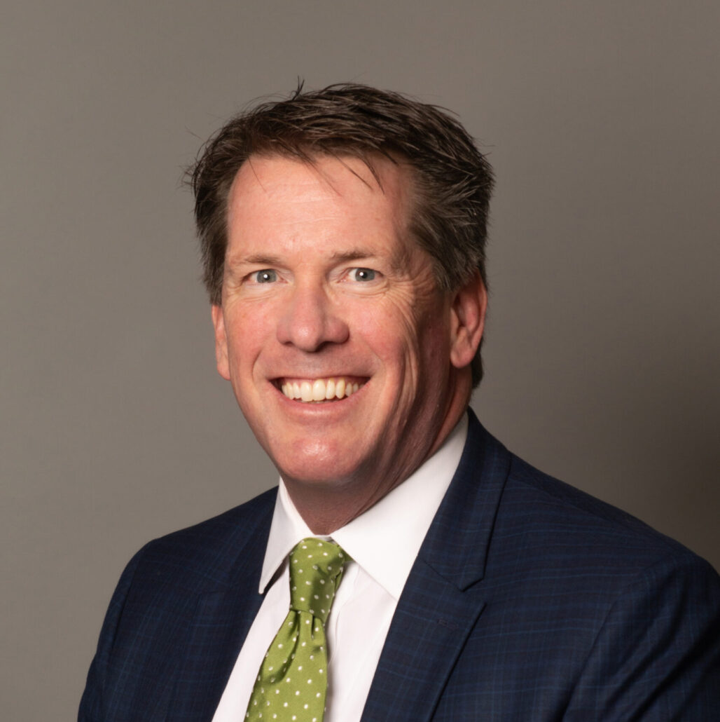 Jim, a white man with brown hair, wears a white shirt with a green tie and black blazer.