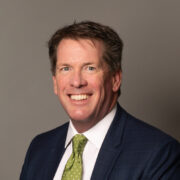 Jim, a white man with brown hair, wears a white shirt with a green tie and black blazer.