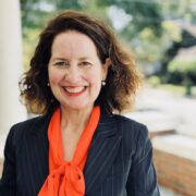Meredith, a white woman with brown hair, wears a bright orange blouse and a black jacket.