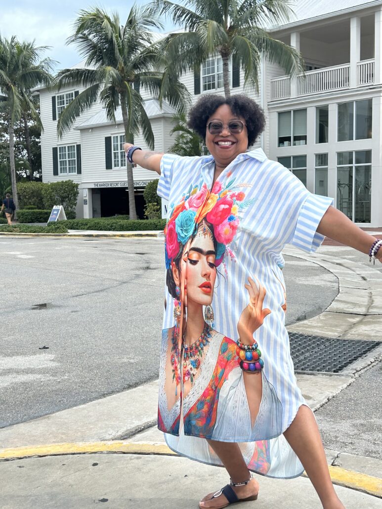 Yolanda, a Black woman with black hair, wears a blue dress and is standing on a street with palm trees behind her.