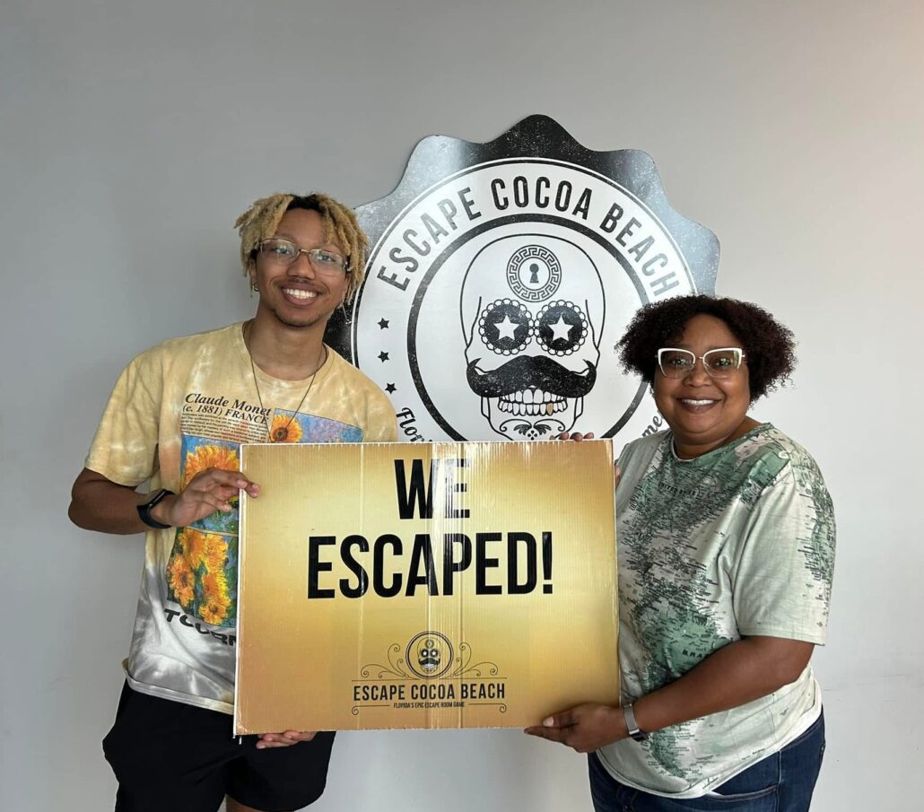 Yolanda, a Black woman with black hair and glasses, wears a pale green shirt. Her son is a Black man with black hair with blond highlights and glasses, and he is wearing a yellow shirt.